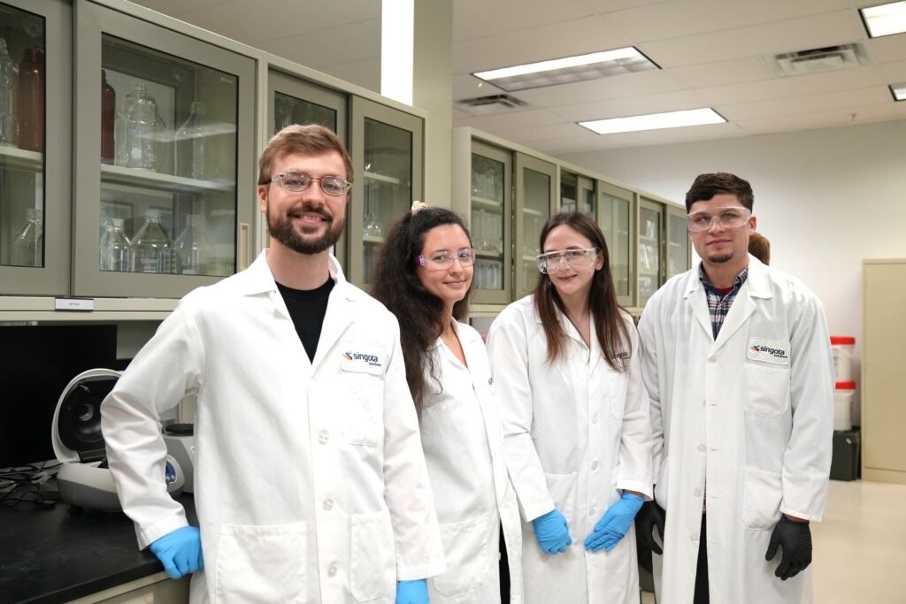Four scientists in white lab coats and safety goggles stand in a laboratory with glass fronted cabinets and lab equipment in the background jun   updated collaborative style jun   updated collaborative style