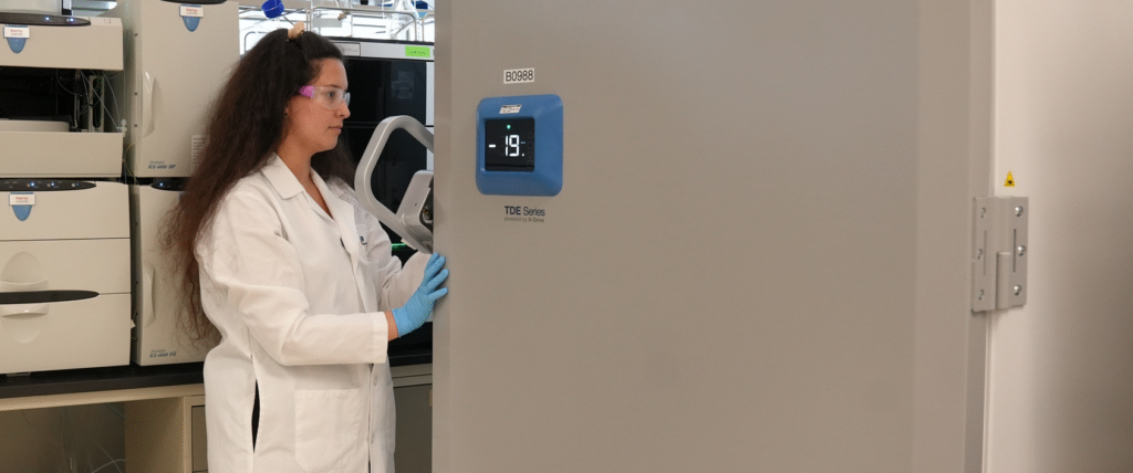Scientist in a white lab coat safety goggles and blue gloves operates a large gray lab freezer watching a blue digital display reading  °C jun updated   ich stability jun updated   ich stability