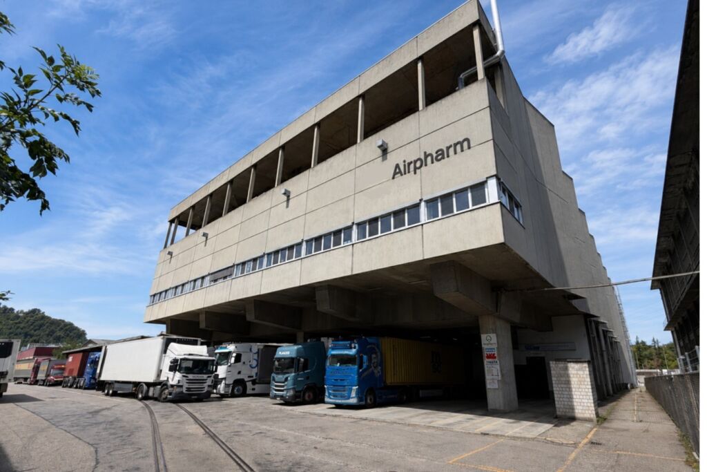Concrete industrial building labeled Airpharm with loading bays and a row of cargo trucks parked beneath the structure under a blue sky basel switzerland custom basel switzerland custom
