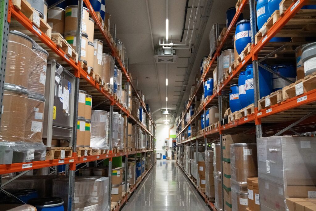 Warehouse aisle with tall orange metal shelving stacked drums and boxes on both sides under fluorescent lights jun updated   basel jun updated   basel