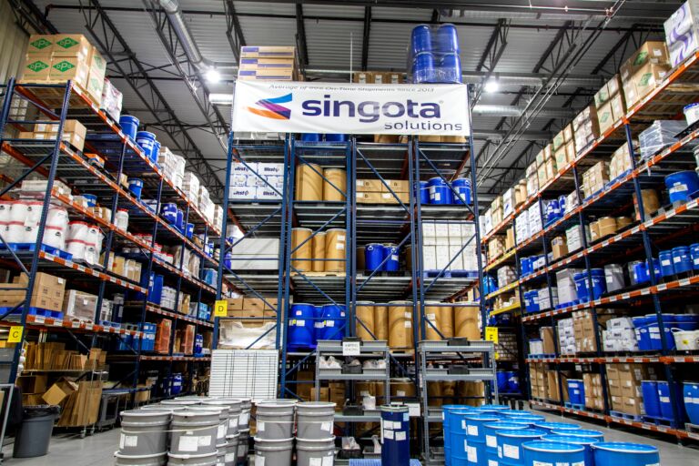 Industrial warehouse with tall blue racking stacked boxes and large blue drums beneath a Singota Solutions banner jun updated   bloomington jun updated   bloomington